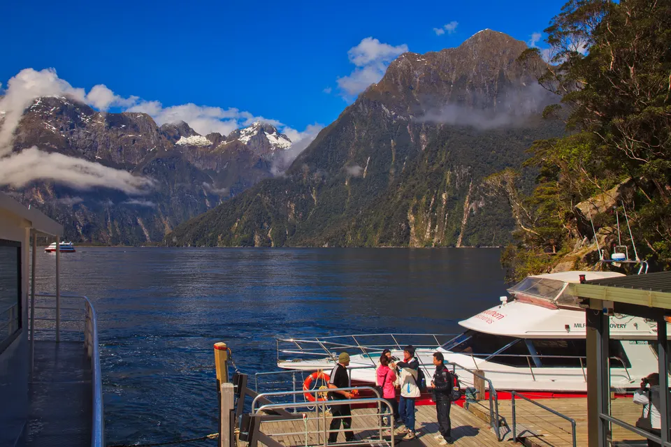 Crucero turístico y kayaks surcando las aguas de espejo bajo los colosales acantilados de Fiordland.