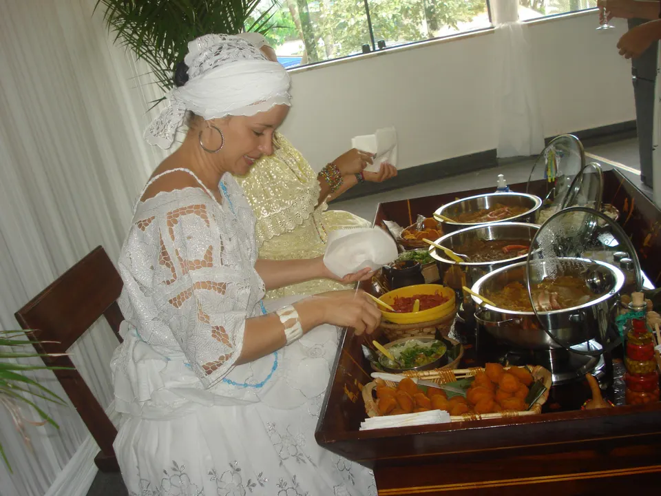 Baiana vestida con traje tradicional preparando acarajé frito en azeite de dendê relleno de vatapá y camarones secos.
